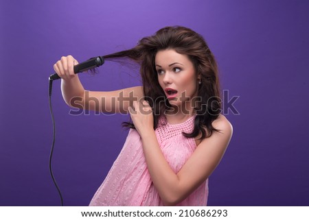 Half-length portrait of astonished young woman with curly hair wearing nice pink dress straightening her hair with the curling irons. Isolated on blue background