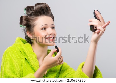 Half-length portrait of beautiful smiling young housewife wearing curlers and green wrapper painting her lips at the mirror. Isolated on blue background