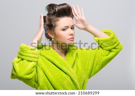 Half-length portrait of pleasant young housewife wearing curlers and green wrapper putting right her haircut. Isolated on blue background