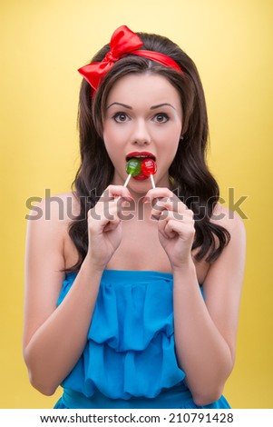 Half-length portrait of funny dark-haired woman wearing nice red headband and blue dress pushing two colorful bonbons in her mouth. Isolated on yellow background