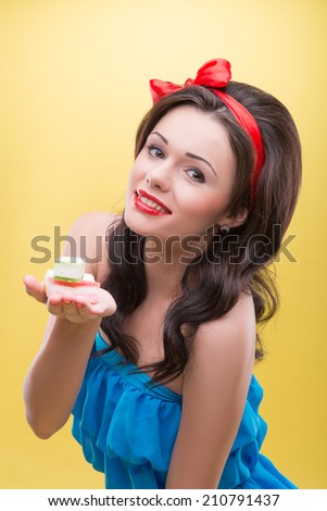 Half-length portrait of charming smiling dark-haired woman wearing nice red headband and wonderful blue dress holding on her palm very delicious colorful fruit drops. Isolated on yellow background