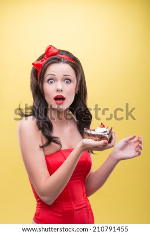 Half-length portrait of very surprised dark-haired woman wearing great red headband holding tasty chocolate cake and looking at us. Isolated on yellow background