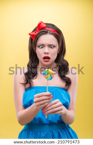 Half-length portrait of pretty dark-haired woman wearing nice red headband and wonderful blue dress looking at her sweet candy in fright. Isolated on yellow background