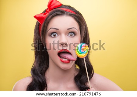 Funny dark-haired girl wearing pretty red headband with wide opened eyes wanted to lick her sweet candy. Isolated on yellow background