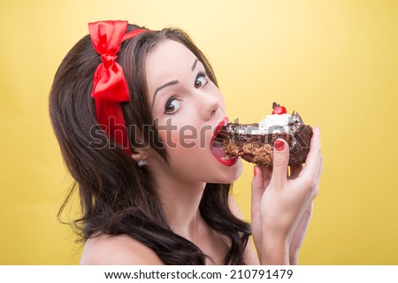 Half-length portrait of very sexy smiling dark-haired woman wearing great red headband biting the piece of tasty chocolate cake and looking at us. Isolated on yellow background