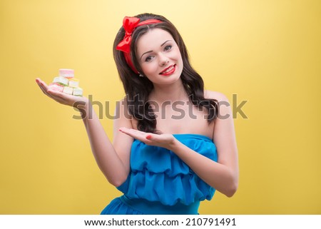 Half-length portrait of charming smiling dark-haired woman wearing nice red headband and wonderful blue dress showing us her favorite delicious colorful fruit drops. Isolated on yellow background
