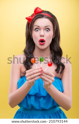Half-length portrait of very excited dark-haired woman wearing nice red headband and blue dress showing us three sweet colorful bonbons. Isolated on yellow background