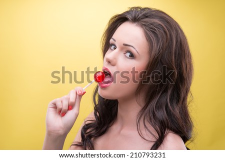 Half-length portrait of sexy dark-haired woman wearing wonderful blue dress standing back to us licking her favorite bonbon. Isolated on yellow background