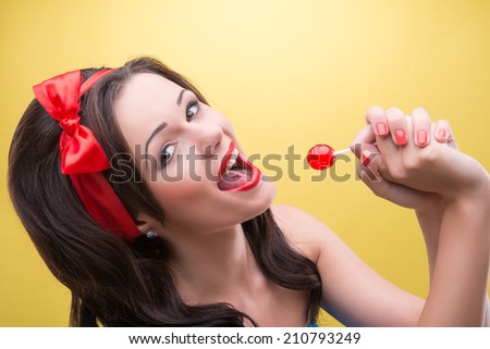 Pretty young smiling dark-haired girl wearing nice red headband wanted to lick very delicious sweet red bonbon. Isolated on yellow background