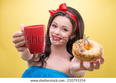 Half-length portrait of lovely smiling dark-haired woman wearing nice red headband and wonderful blue dress suggested us her aromatic coffee and tasty doughnut. Isolated on yellow background