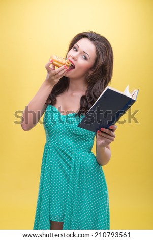 Half-length portrait of very beautiful dark-haired woman relishing her favorite sweet doughnut reading the interesting book. Isolated on yellow background