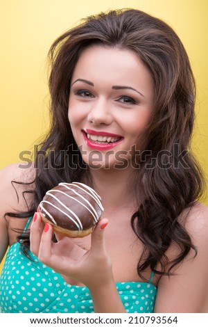 Half-length portrait of very beautiful smiling dark-haired woman wearing nice mint dotted dress wanted to try her appetizing round doughnut covered with the chocolate icing. Isolated on yellow