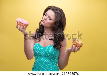 Half-length portrait of pretty dark-haired woman wearing nice mint dotted dress cannot to choose the tastiest doughnut. Isolated on yellow background