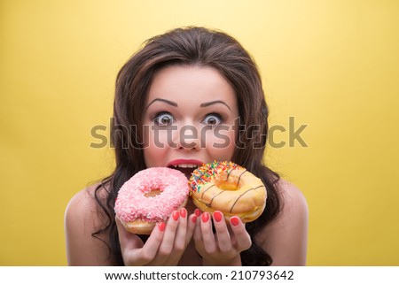 Half-length portrait of very excited dark-haired woman wearing nice blue dotted dress taking delight in very delicious doughnuts. Isolated on yellow background