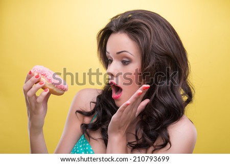 Very beautiful dark-haired woman with great make up surprised with the size of her doughnut covered with pink icing. Isolated on yellow background