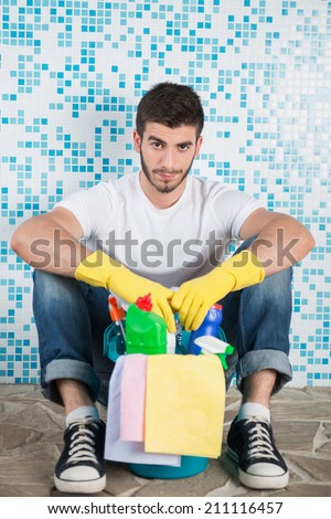 Young dark-haired janitor wearing white shirt blue jeans and yellow rubber gloves sitting in the bathroom with pail of cleaners leaning on the wall