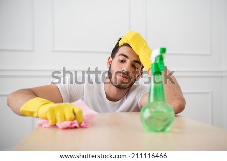 Half-length portrait of young dark-haired janitor wearing white shirt and yellow rubber gloves dusting the table very diligently