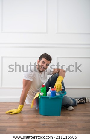 Young smiling dark-haired janitor wearing white shirt blue jeans and yellow rubber gloves sitting on the floor in the office near his pail of cleaners having a rest