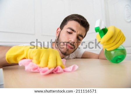 Half-length portrait of young dark-haired janitor wearing white shirt and yellow rubber gloves dusting the table very carefully
