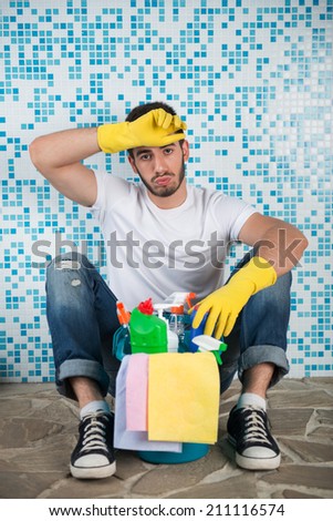 Young tired dark-haired janitor wearing white shirt blue jeans and yellow rubber gloves sitting in the bathroom with pail of cleaners leaning on the wall and wiping her head with the hand