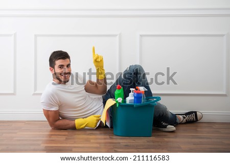 Young smiling dark-haired janitor wearing white shirt blue jeans and yellow rubber gloves lying on the floor near pail of cleaners relaxing