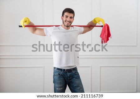 Half-length portrait of young smiling dark-haired janitor wearing white shirt blue torn jeans and yellow rubber gloves standing with the red mop on his shoulders in the room