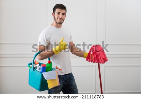 Half-length portrait of young dark-haired janitor wearing white shirt blue torn jeans and yellow rubber gloves standing with the red mop in one hand and pail with cleansers in another showing