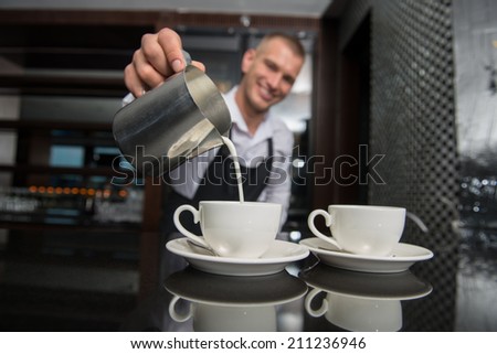 Selective focus on two white cups of coffee and metal jar with milk. Handsome young smiling barista wearing white shirt and black apron pouring the milk on background