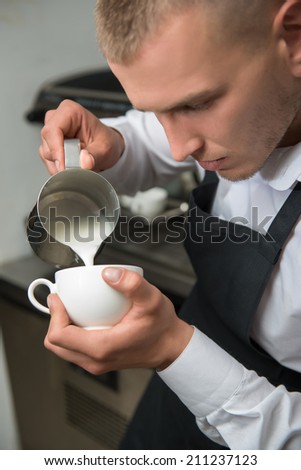 Half-length portrait of handsome young concentrated barista wearing white shirt and black apron pouring the milk into the cup of coffee