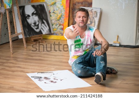 Fair-haired handsome painter wearing painted white shirt and blue torn jeans sitting on the floor in his workshop among the pictures and inviting us to him