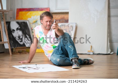 Fair-haired handsome painter wearing painted white shirt and blue torn jeans sitting on the floor in his workshop among the pictures and looking very attentively at his lead pencil