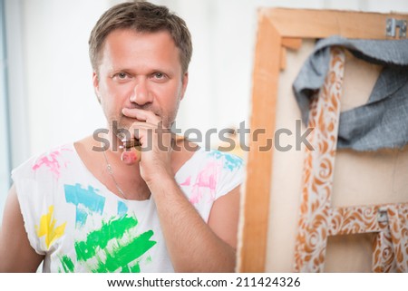 Half-length portrait of fair-haired thoughtful painter wearing white painted shirt standing near his picture holding many brushes in his hand and looking at us
