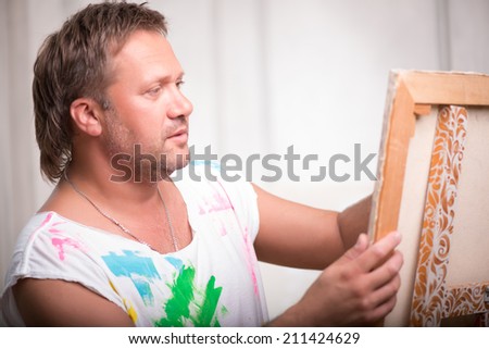 Half-length portrait of fair-haired mature painter wearing white painted shirt holding his picture and looking at it