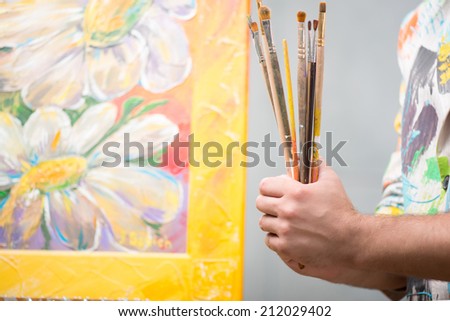 Painter wearing white painted jacket holding a pile of different brushes in his hand standing near his lovely picture