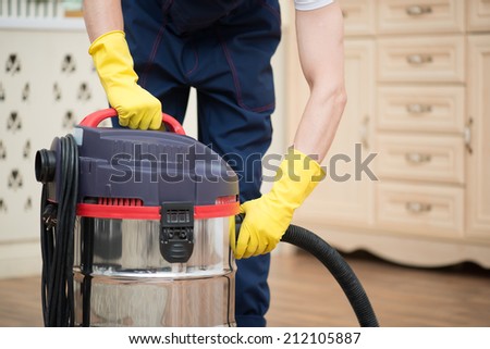 Selective focus on janitor wearing white T-shirt blue overalls and yellow rubber gloves turning off the vacuum cleaner. His working place on background