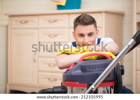 Selective focus on dark-haired smiling janitor wearing white T-shirt blue overalls and yellow rubber gloves leaning on vacuum cleaner. Commode on background