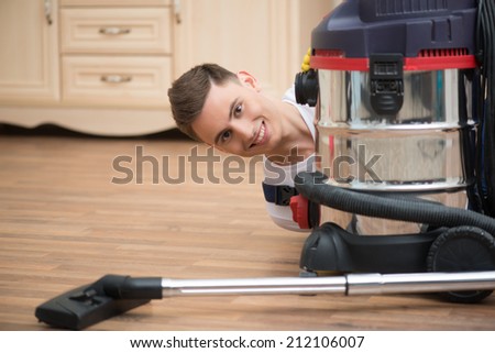 Selective focus on dark-haired smiling janitor wearing white T-shirt blue overalls and yellow rubber gloves looking out of the vacuum cleaner. His working place on background