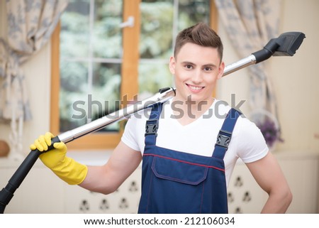 Selective focus on dark-haired smiling janitor wearing white T-shirt blue overalls and yellow rubber gloves standing with the hose of vacuum cleaner on his shoulder. Window on background