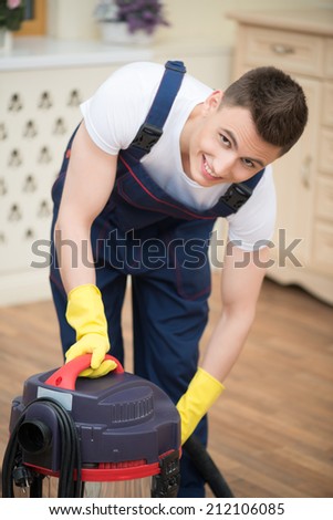 Selective focus on dark-haired smiling janitor wearing white T-shirt blue overalls and yellow rubber gloves turning off the vacuum cleaner. His working place on background
