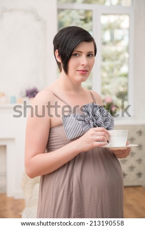 Selected focus on dark-haired lovely pregnant woman wearing great long dress staying drinking a tea. Her living room on background