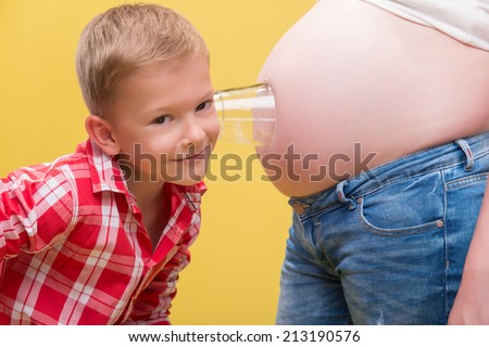 Half-length portrait of pregnant woman wearing white T-shirt and jeans standing with her smiling little son wearing red checked shirt trying to hear the baby through the glass. Isolated on yellow