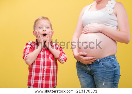 Half-length portrait of pregnant woman wearing white T-shirt and jeans standing with her scared little son does not know what happened to his mom. Isolated on yellow background