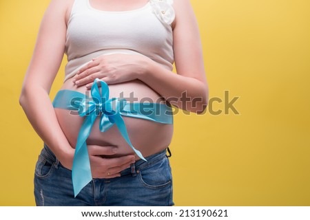 Half-length portrait of pregnant woman wearing white T-shirt and jeans having a blue band on her belly dreaming about her baby. Isolated on yellow background