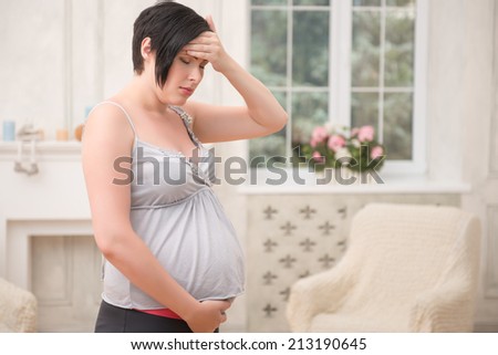 Selected focus on dark-haired anxious pregnant woman wearing nice shirt and black pants standing touching her belly having a headache. Her living room on background