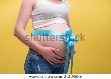 Half-length portrait of pregnant woman wearing white T-shirt and jeans standing aside with blue band on her belly dreaming about her baby. Isolated on yellow background