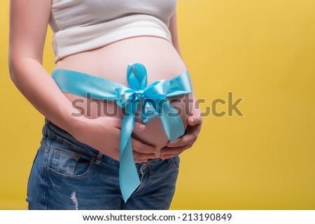 Half-length portrait of pregnant woman wearing white T-shirt and jeans standing aside with blue band on her belly dreaming about her baby. Isolated on yellow background