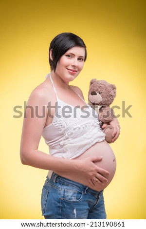 Half-length portrait of young pretty dark-haired smiling pregnant woman wearing white T-shirt and jeans holding teddy bear.