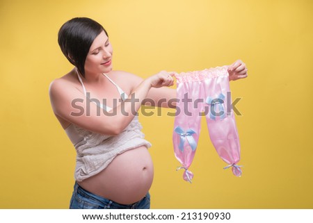 Half-length portrait of young pretty dark-haired smiling pregnant woman wearing white T-shirt and jeans standing with small pink pants for baby girl looking at them. Isolated on yellow background