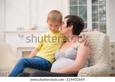 Selected focus on dark-haired lovely pregnant woman wearing nice shirt and black pants sitting in the comfortable chair with her little son wanted to say him something secretly. Her living room on