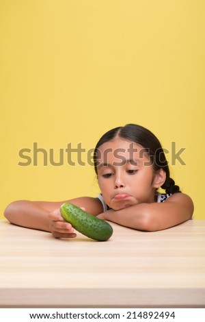 Half-length portrait of pretty dark-haired sad little girl wearing nice striped dress sitting at the table and dolefully looking at the cucumber. Isolated on yellow background